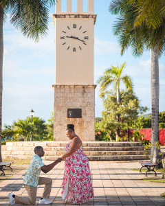 Romantic marriage proposal at the iconic Clock Tower in Frederiksted, St. Croix — a man kneeling with a ring while his emotional partner covers her mouth in surprise, surrounded by the vibrant harbor and historic architecture of the US Virgin Islands