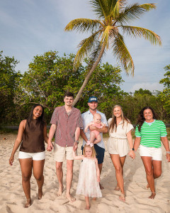 Family smiling together on a tropical beach with turquoise ocean and palm trees in the background.
