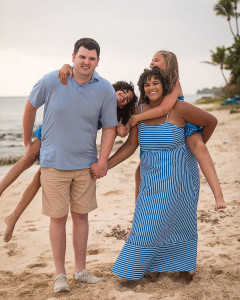 Family of four smiling together on a tropical beach with turquoise ocean and palm trees in the background.