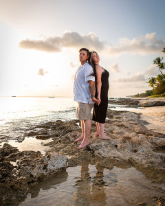 Couple standing close together on the beach at sunset, facing the ocean with soft golden light around them.